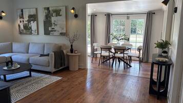 A spacious living room with a sofa, coffee table, and dining area with chairs and a table. There are abstract paintings on the wall, a vase with branches, and a potted plant on a side table.