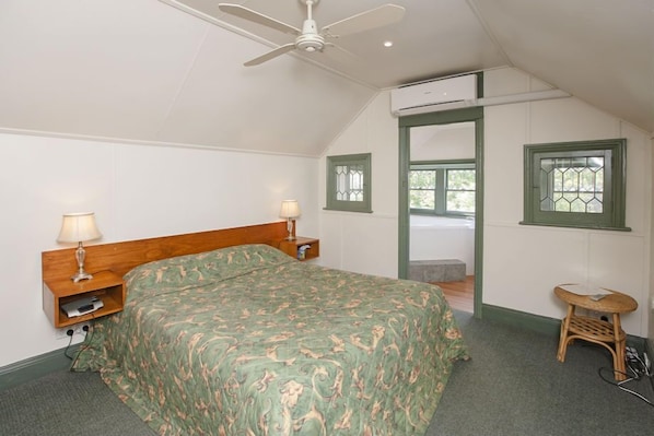 A bedroom with a bed, two bedside tables with lamps, a ceiling fan, a window with green trim, and a small stool.