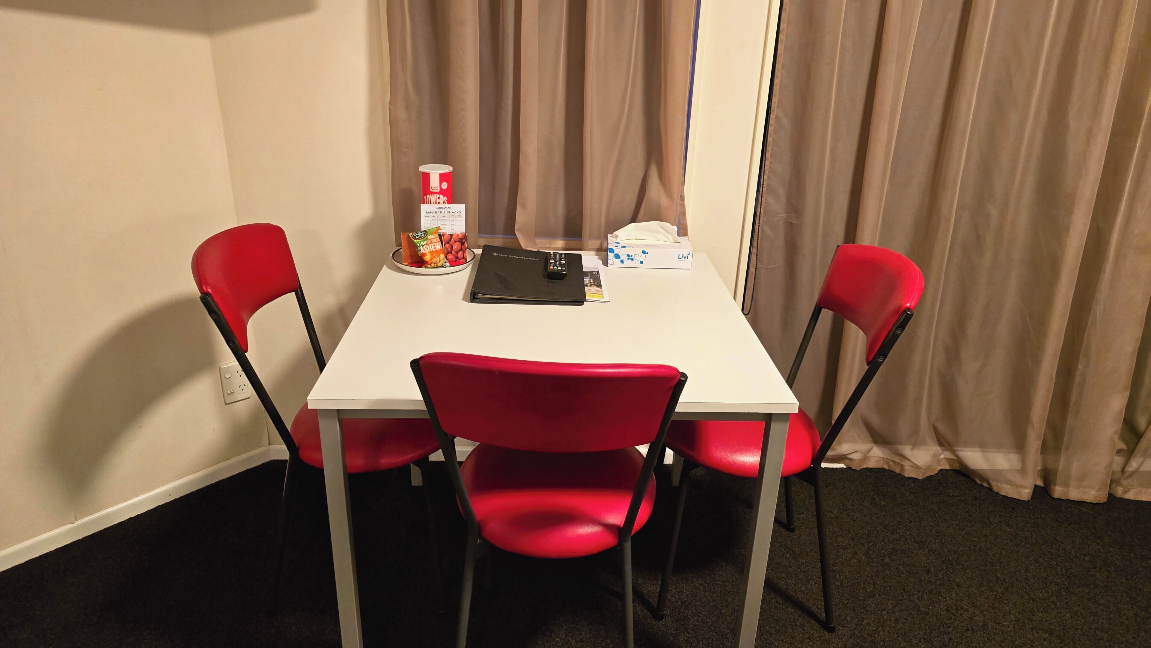 Une petite chambre d’hôtel avec une table blanche, deux chaises rouges, un classeur noir, une boîte de mouchoirs et une boîte de noix.