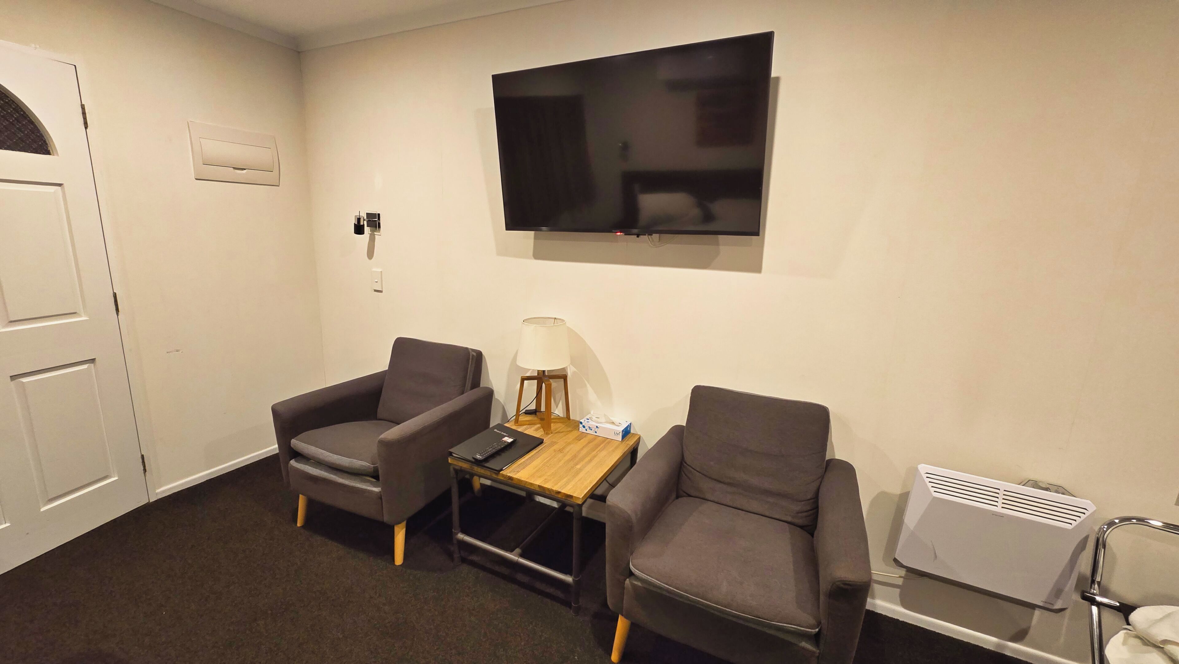 Una habitación con dos sillones, una mesita con lámpara, un televisor montado en la pared y un calentador en el suelo.