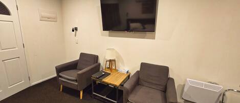 Una habitación con dos sillones, una mesita con lámpara, un televisor montado en la pared y un calentador en el suelo.