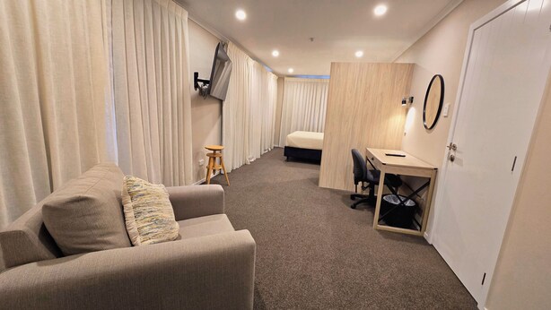 A hotel room with a sofa, a desk, a chair, a bed, and a wardrobe. - Carramar Motor Inn (Palmerston North)