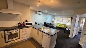 A compact kitchen with a microwave, kettle, and stove, a dishwasher, and a dining area with a sofa and a small table. - Carramar Motor Inn (Palmerston North)