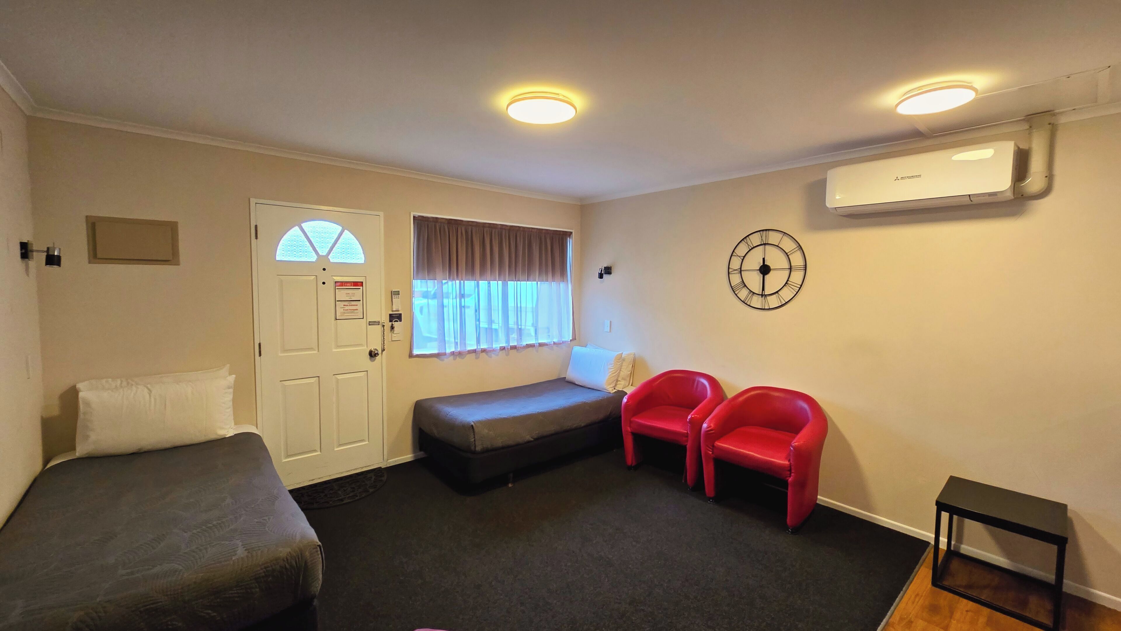 A hotel room with two single beds, a sofa bed, two red chairs, a clock, and an air conditioning unit.