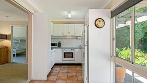 A compact kitchen with white cabinets, a built-in oven, and a microwave. There's a clock on the wall and a window with a view of greenery outside. - Glen Isla House (Cowes)