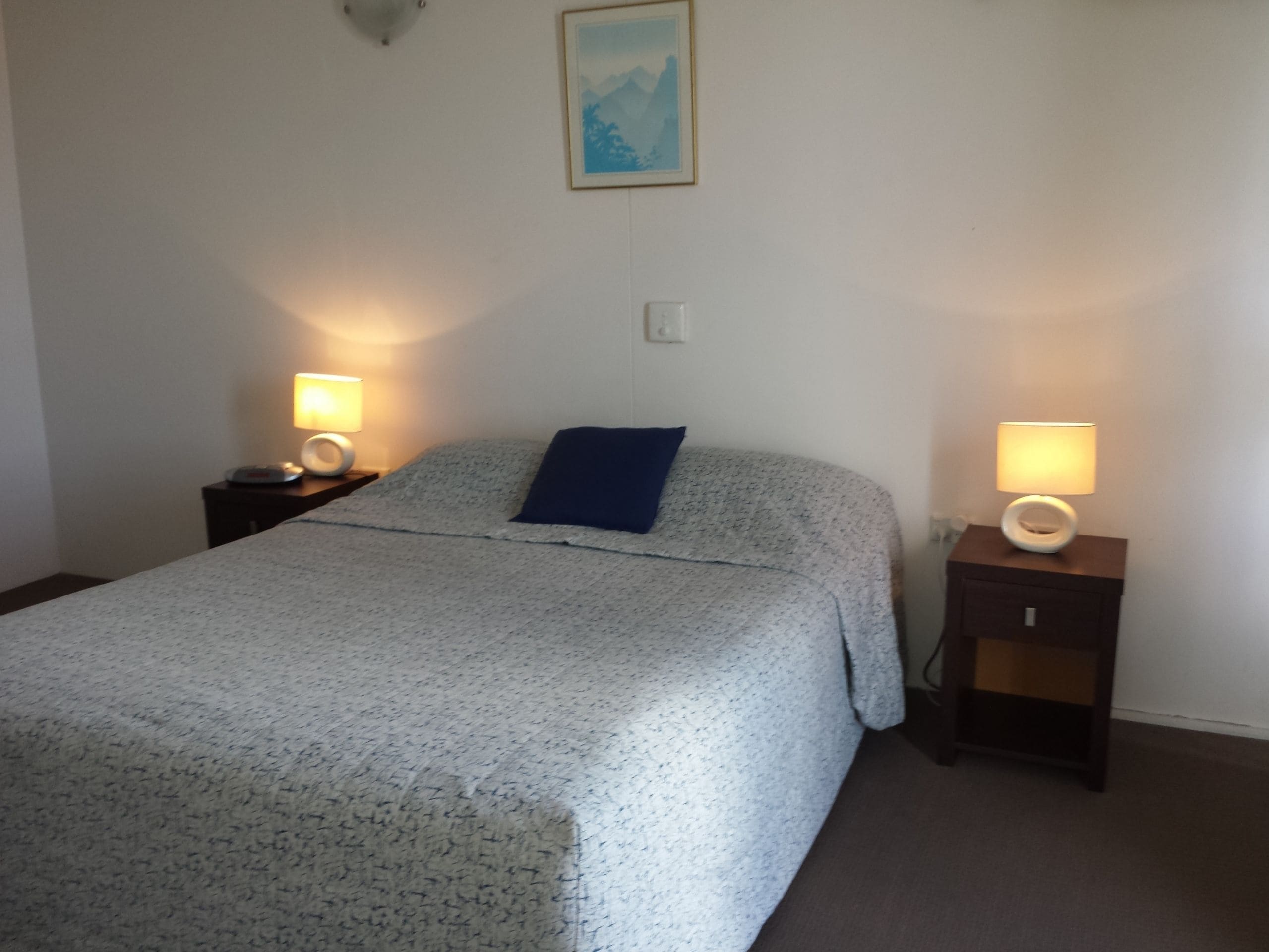 A bedroom with a bed, two bedside tables with lamps, a wall-mounted light fixture, and a framed picture on the wall.