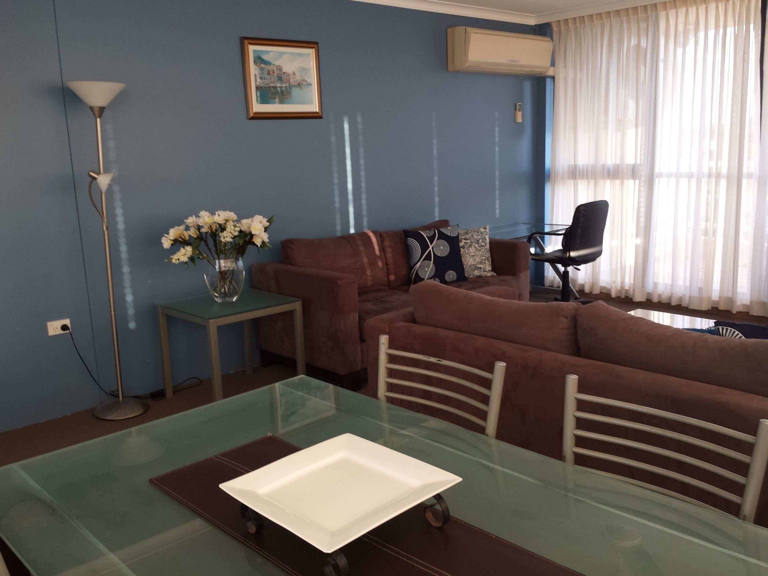 Una sala de estar con una mesa de comedor de vidrio, un sofá marrón y un cuadro en la pared.