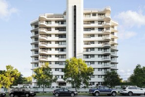 A multi-story residential building with balconies, a central vertical feature, and parked cars in front. - Kirribilli Riverfront Apartments (New Farm)