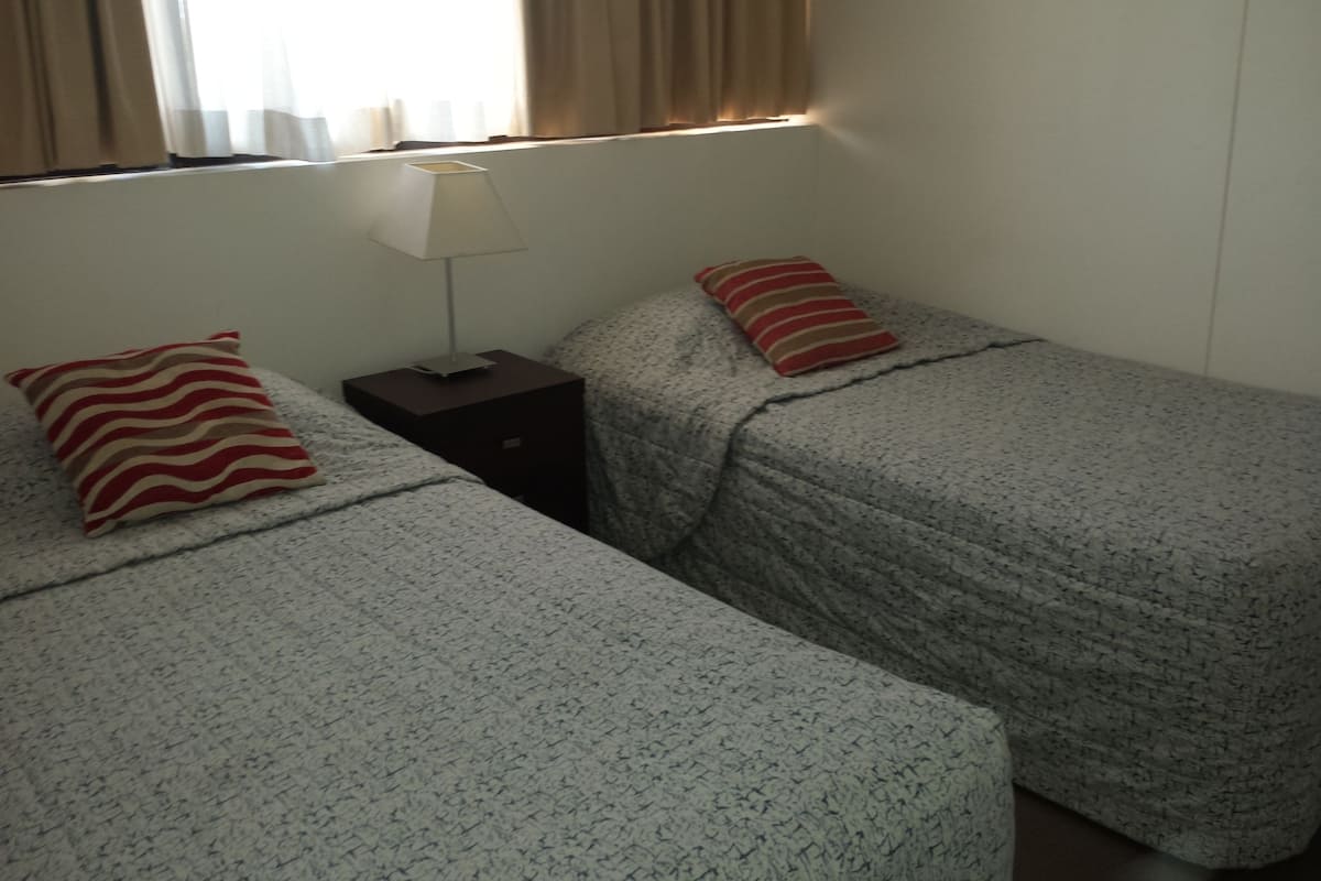 A room with two beds, each with a patterned bedspread and a striped pillow. There is a nightstand with a lamp between the beds.