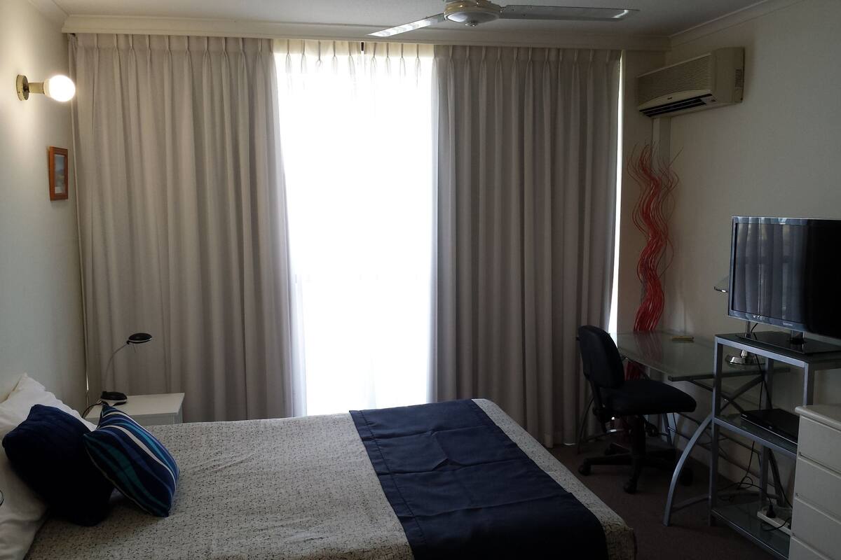 A hotel room with a bed, a desk with a chair, a television, an air conditioner, and a window with curtains.