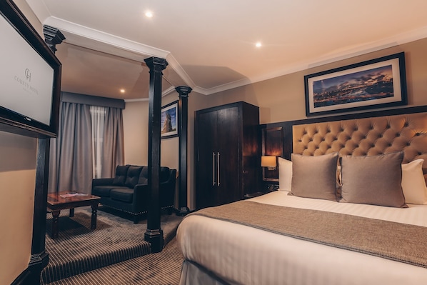 A hotel room with a large bed, a dark wooden wardrobe, a dark armchair, a small table, and a framed picture on the wall.