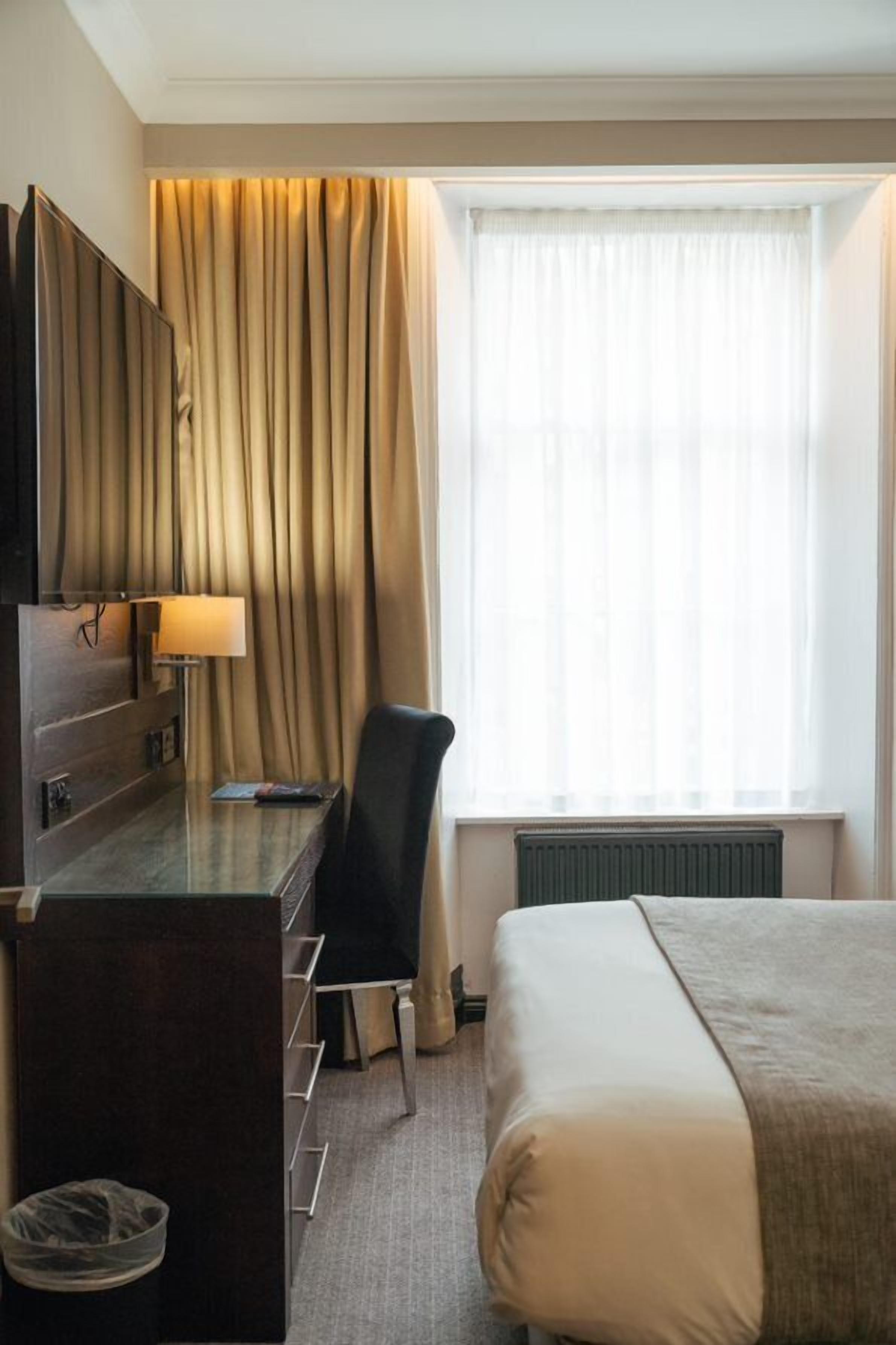 A hotel room with a bed, a desk with a lamp, a chair, a TV, and a window with curtains.