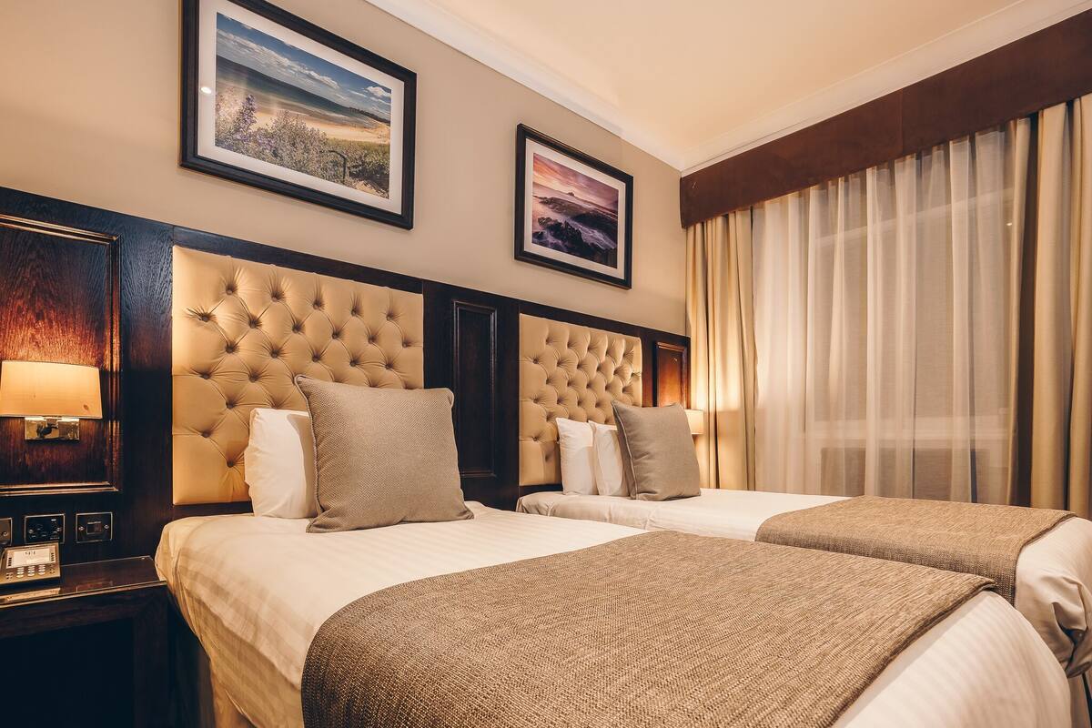 A hotel room with two beds, a large window with curtains, a wall-mounted lamp, and framed pictures.