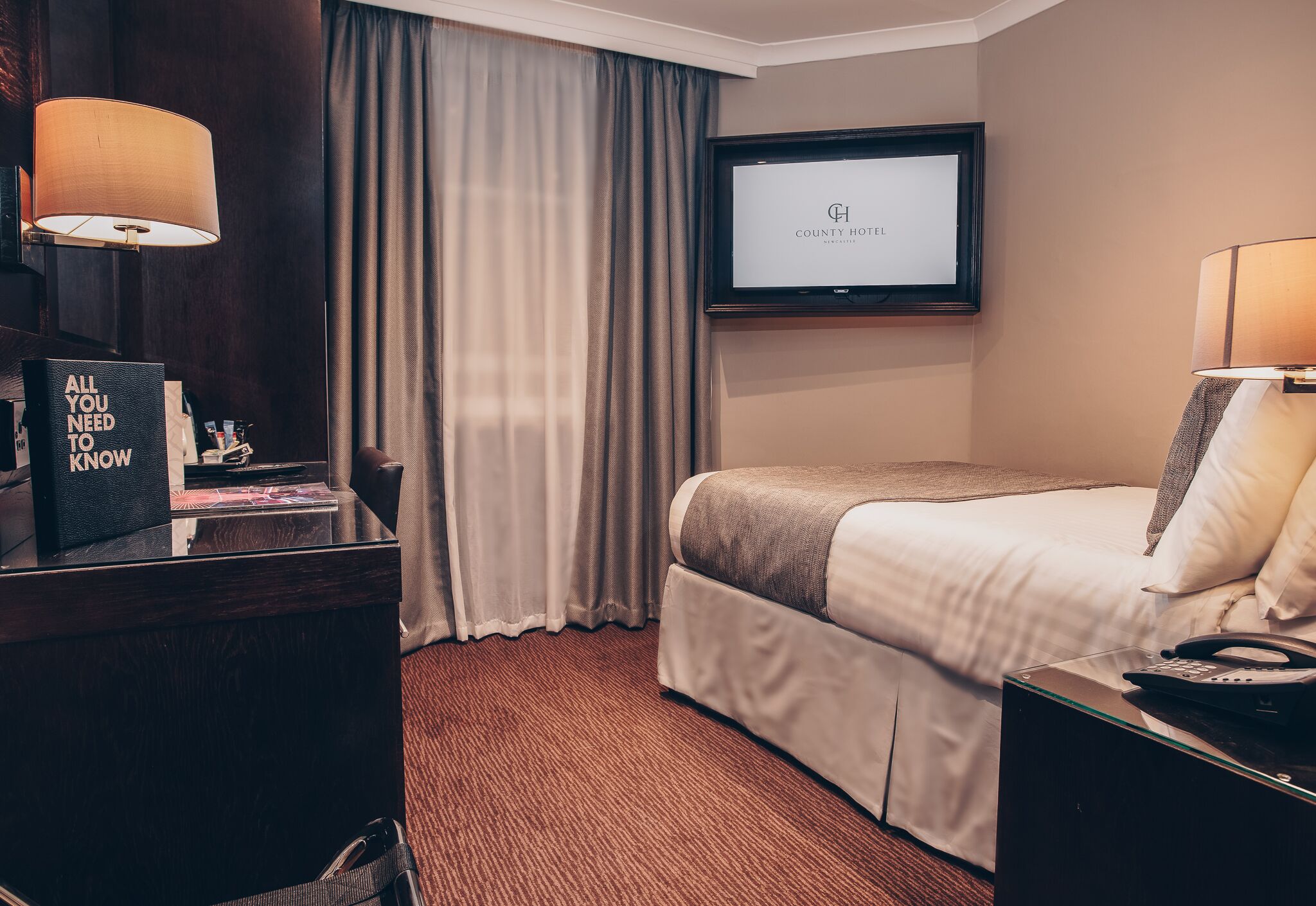 Quarto de hotel com cama, escrivaninha com luminária, televisão na parede e placa com a mensagem 'Tudo que você precisa saber'.