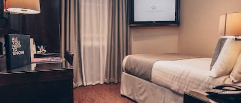 Quarto de hotel com cama, escrivaninha com luminária, televisão na parede e placa com a mensagem 'Tudo que você precisa saber'.