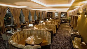 Ein Restaurant mit runden Tischen, gepolsterten Sitzgruppen, Teppichböden und Vorhängen an den Fenstern.