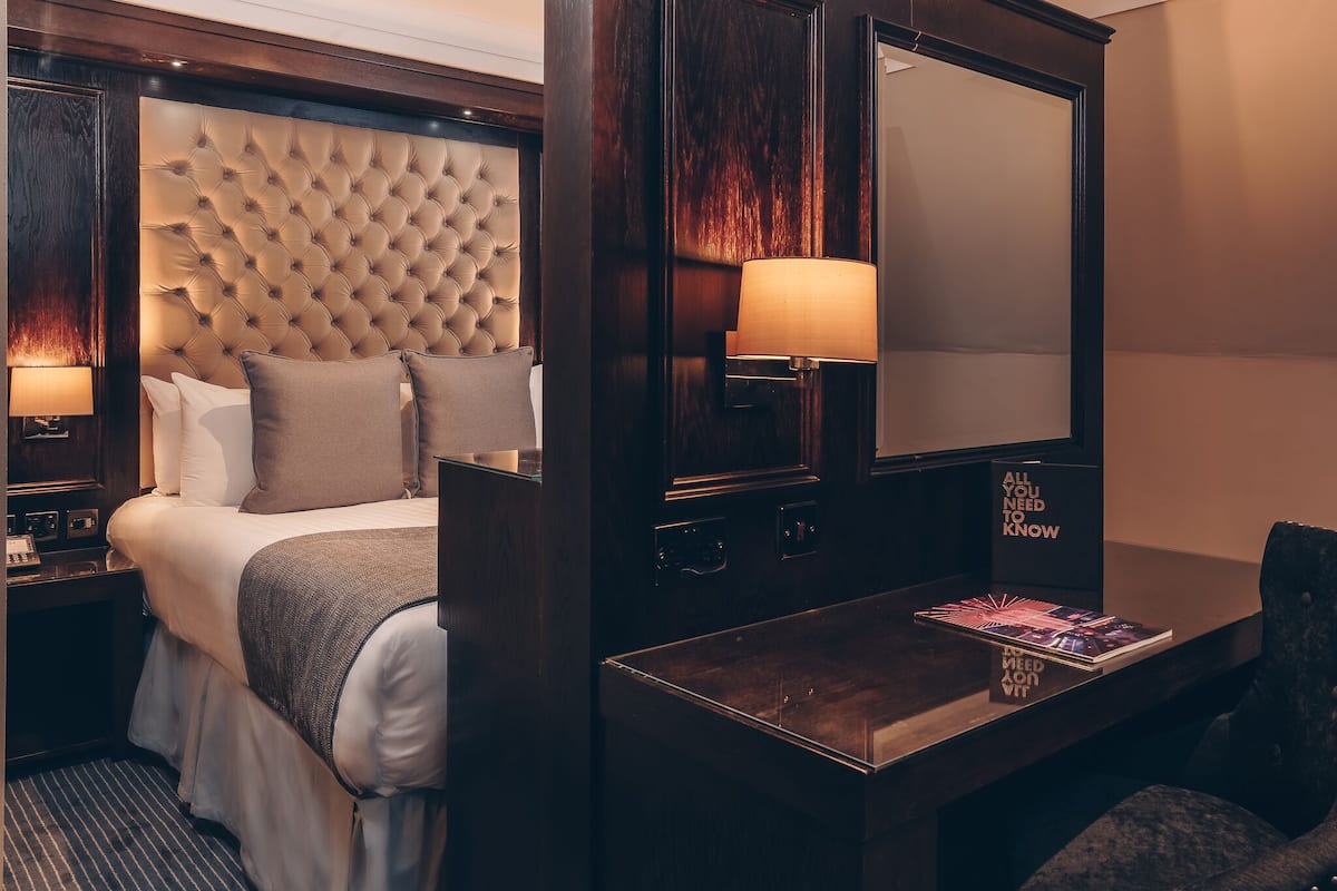 A hotel room with a tufted headboard, a bedside table with a magazine, and a large mirror.