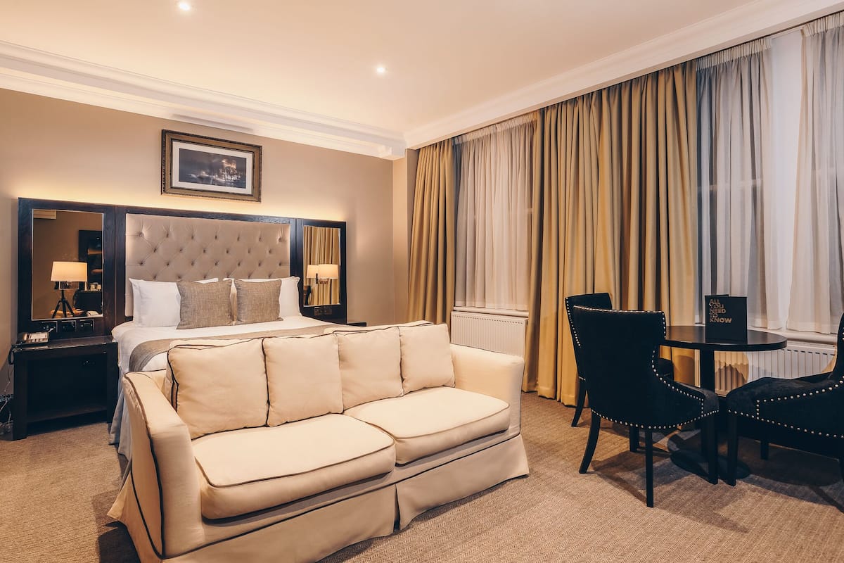 A hotel room with a large bed, a sofa, a dining table with chairs, a mirror, and a window with curtains.