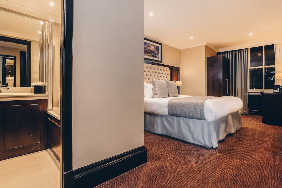 A hotel room with a large bed, a bedside table, a mirror, a window with curtains, and a framed picture on the wall.