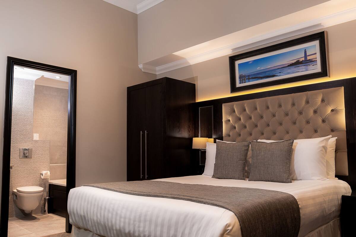 A modern hotel room with a large bed, a headboard with tufting, a framed picture above the bed, and a dark wardrobe.