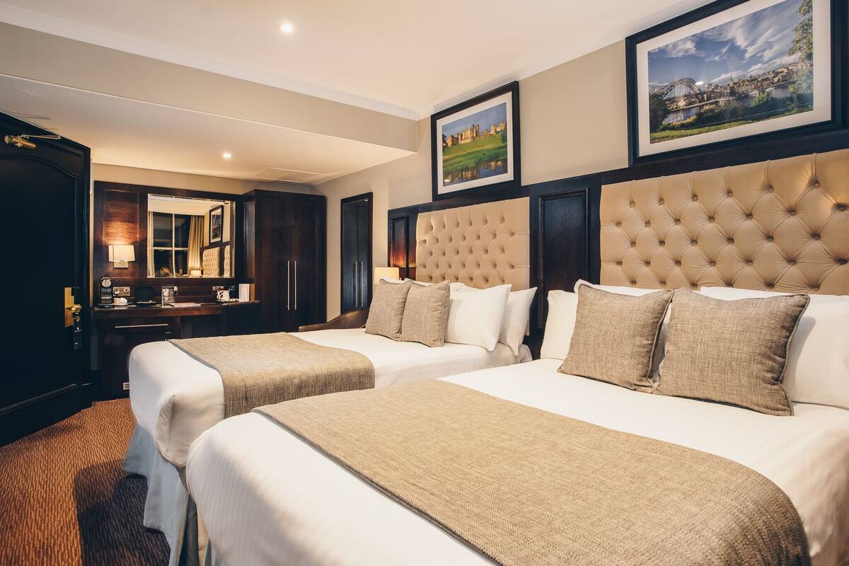 A hotel room with two beds, a large window, a desk with a lamp, and framed pictures on the wall.