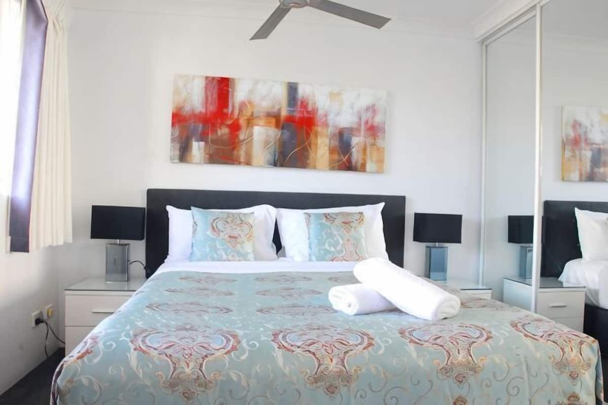 A hotel room with a bed, two bedside tables with lamps, a ceiling fan, and a large mirror.