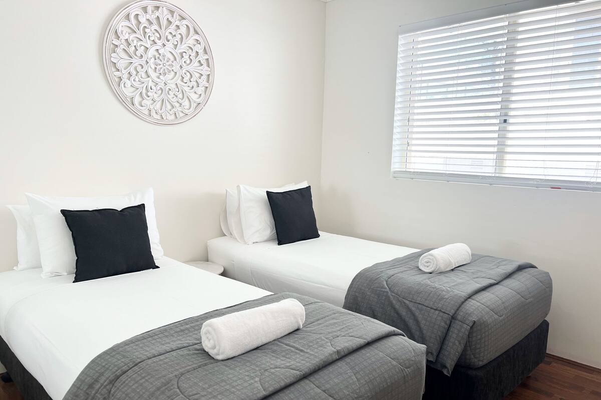 A hotel room with two single beds, each with a grey quilt and a rolled white towel. There is a decorative wall piece above the beds and a window with blinds to the right.