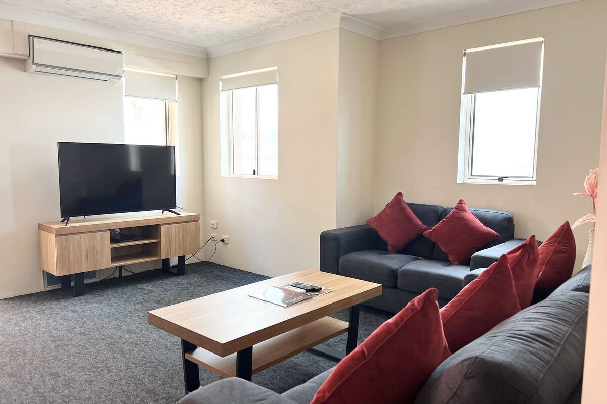 A modern living room with a flat-screen TV, a wooden entertainment unit, a grey sofa with red cushions, a wooden coffee table, and two windows providing natural light.