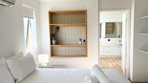 A neatly made bed with white linens and a rolled towel, a bedside lamp, and a wooden shelf with a plant and two electronic devices. - Montego Sands Resort (Mermaid Beach)