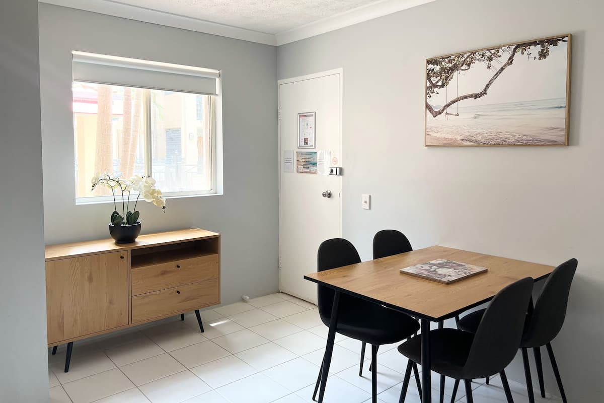 A room with a wooden desk, a window with curtains, a door with a poster, a painting on the wall, and a tiled floor.