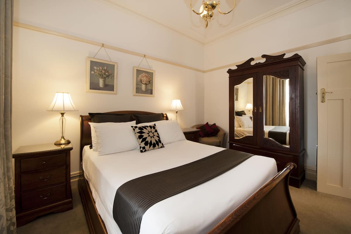 A neatly made bed with white linens and a black throw, flanked by two bedside tables with lamps, a large mirror, and two framed floral paintings on the wall.