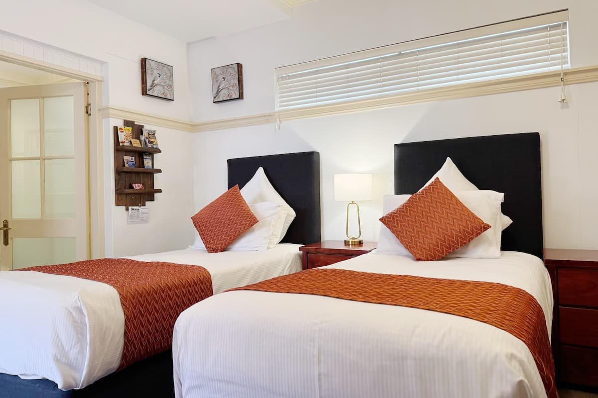A hotel room with two beds, each with a black headboard, white bedding, and orange patterned pillows. There is a small bedside table with a lamp, a shelf with items, and a window with blinds.