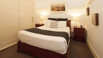 A neatly made bed with white linens and a dark throw blanket, flanked by two bedside tables with lamps, in a room with a large painting on the wall and a door to the left.