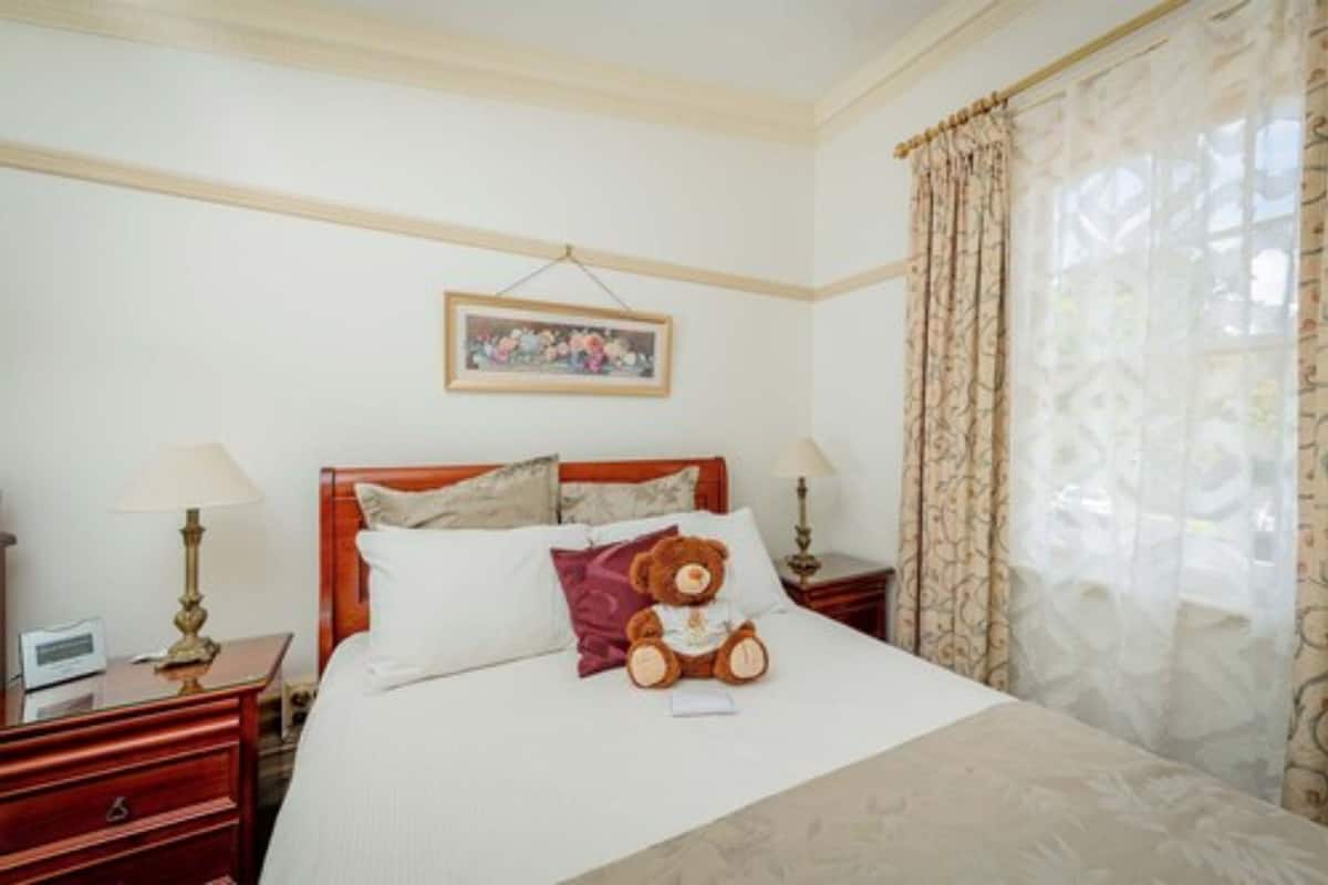 A hotel room with a bed, a nightstand, a lamp, a teddy bear, and a painting on the wall.