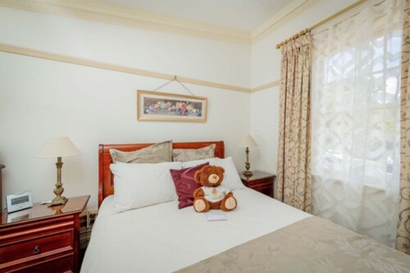 A hotel room with a bed, a nightstand, a lamp, a teddy bear, and a painting on the wall. - Cobb & Co Court Boutique Hotel (Mudgee)