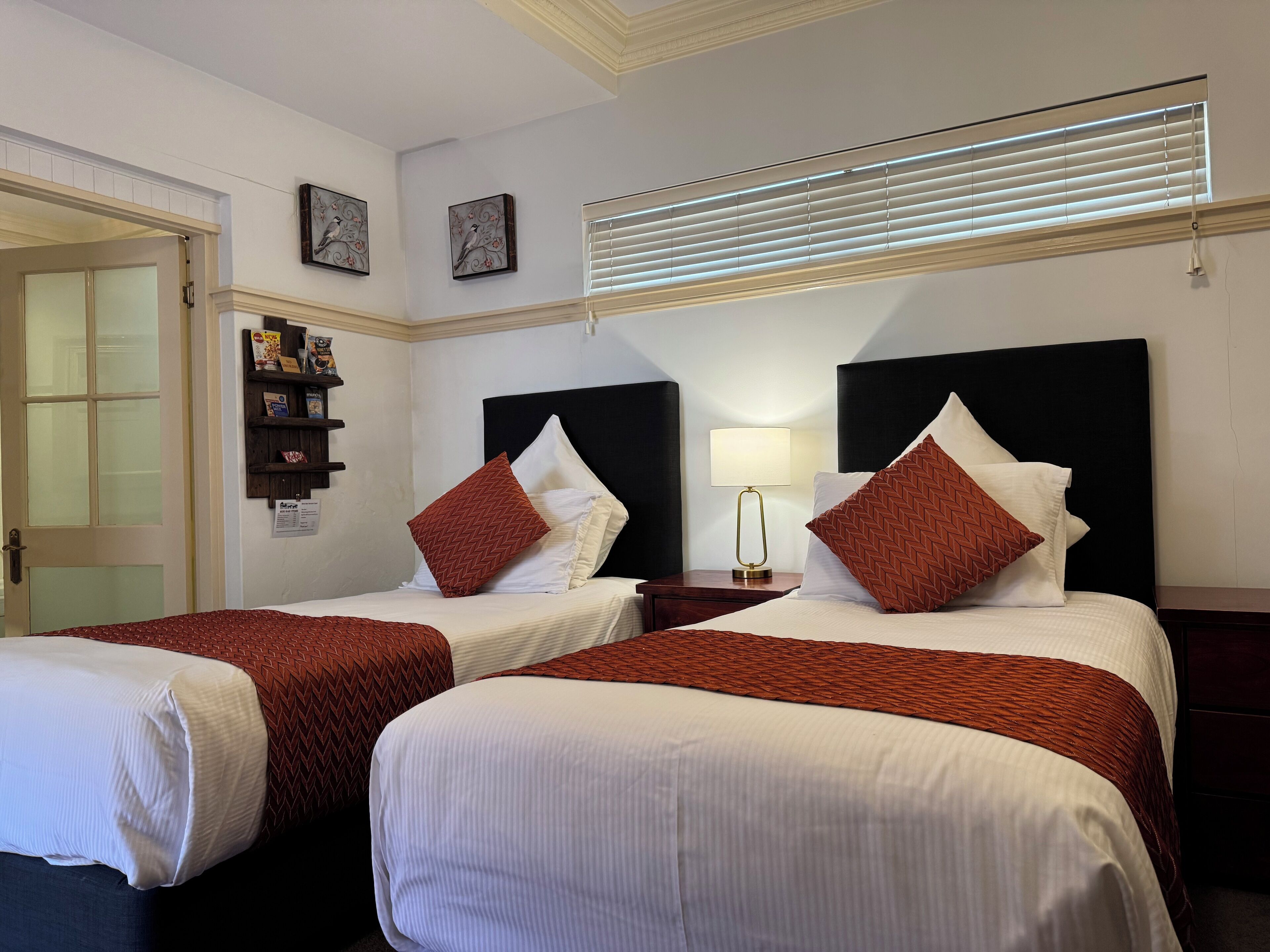 Habitación de hotel con dos camas, cada una con ropa de cama blanca y almohadas rojas estampadas. Hay una mesita de noche con una lámpara entre las camas. La habitación tiene una puerta de madera, un estante con libros y un pequeño reloj en la pared, y una ventana con persianas.