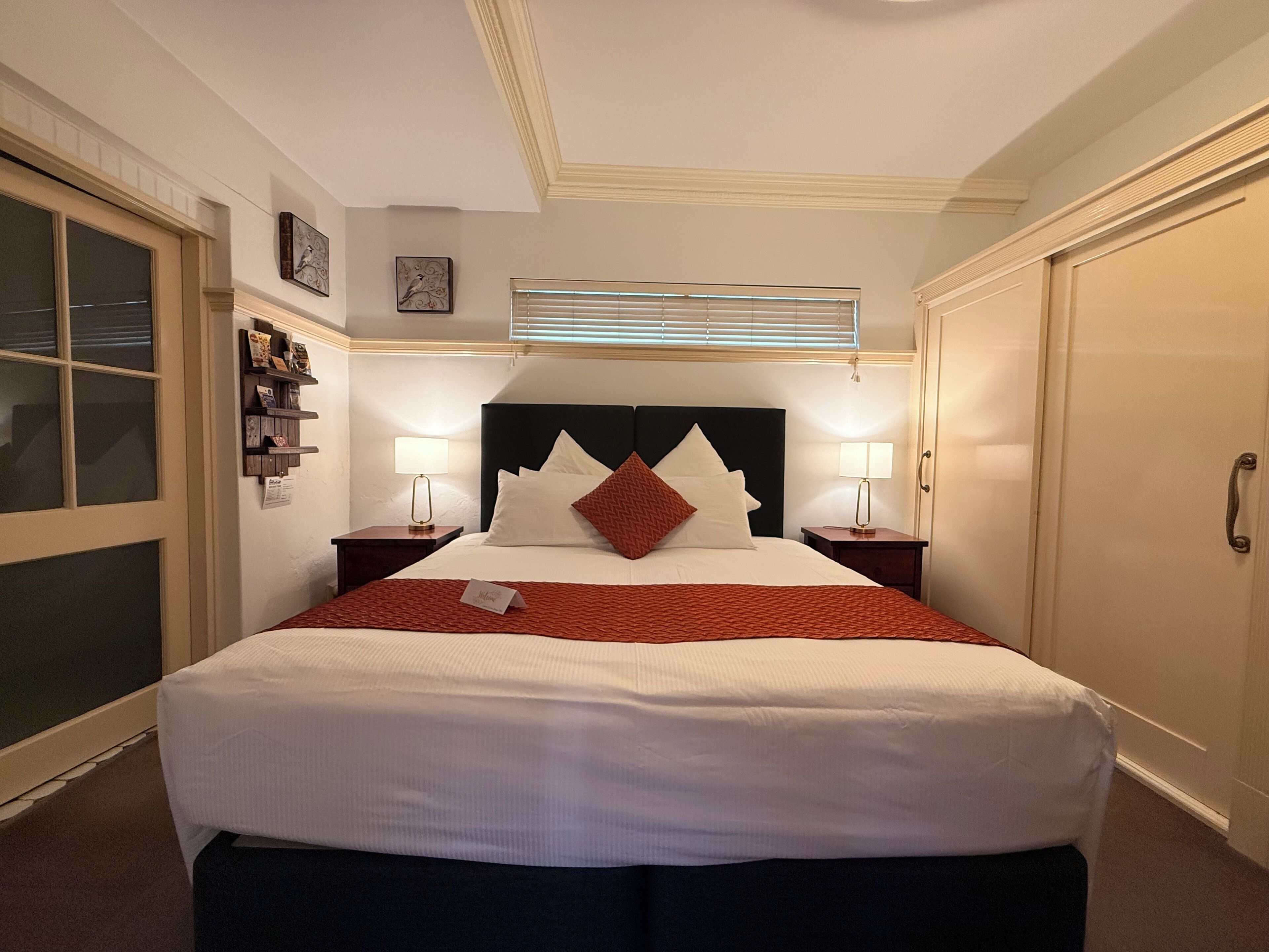 A neatly arranged hotel room with a large bed, two bedside tables with lamps, a window with blinds, and a door.