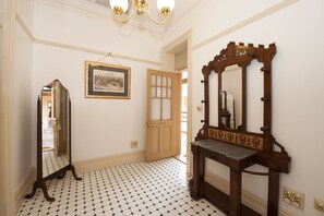Um corredor com piso de azulejos, porta de madeira, quadro decorativo na parede e um espelho grande com moldura de madeira.
