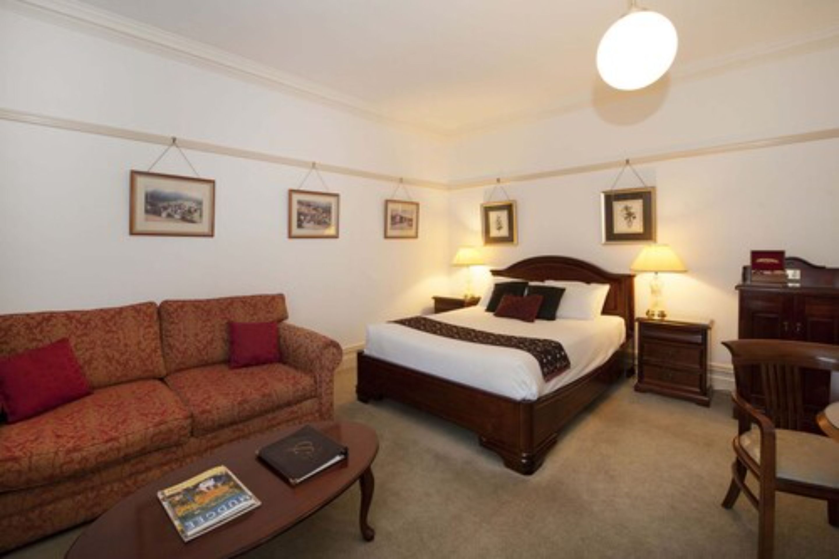 A hotel room with a bed, a sofa, a small table, and a chair. There are framed pictures on the wall and a lamp on the bedside table.