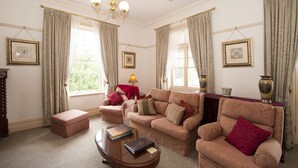 A living room with two sofas, a coffee table, and a red ottoman. There are curtains, framed pictures on the walls, and a chandelier. - Cobb & Co Court Boutique Hotel (Mudgee)