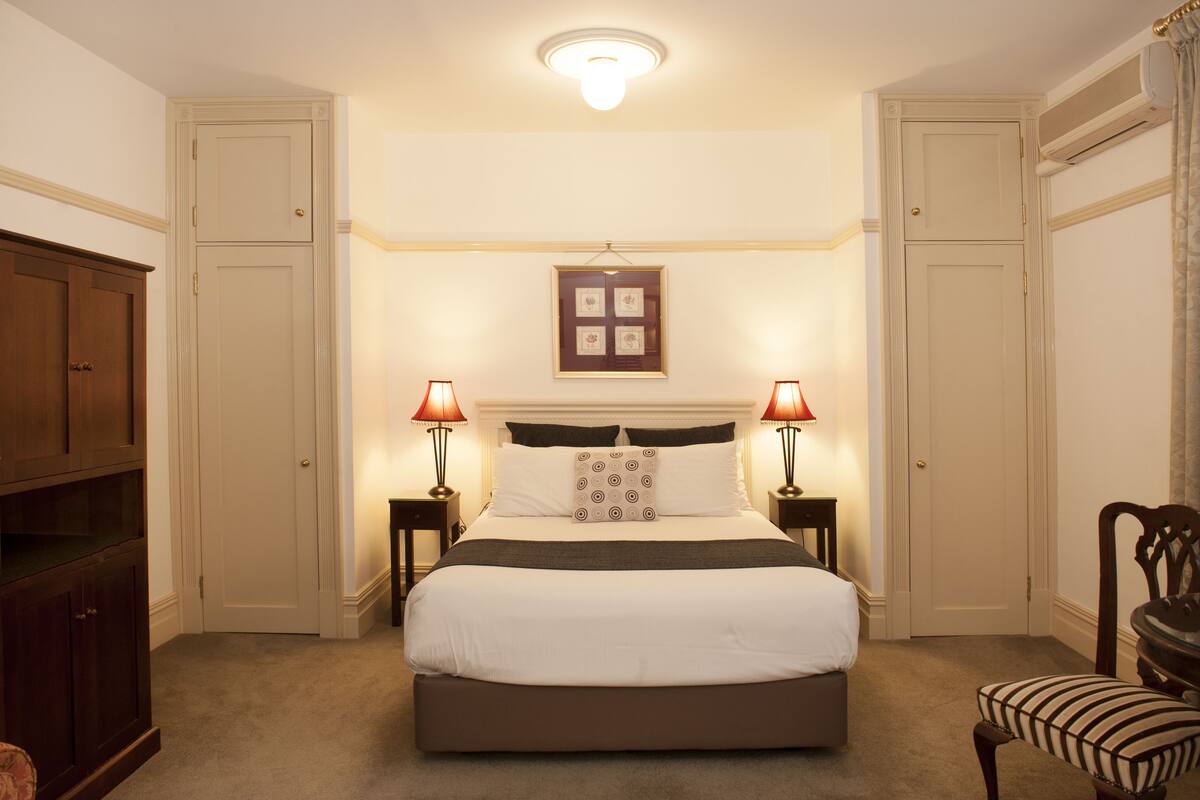A neatly arranged bedroom with a large bed, two bedside tables with lamps, a framed picture above the bed, and a wardrobe on one side.