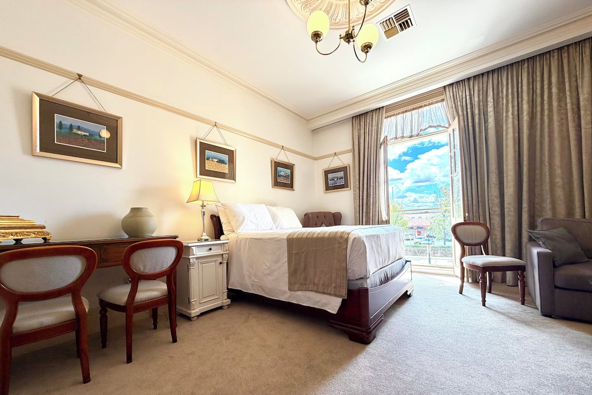 A hotel room with a bed, two chairs, a desk with a lamp, and framed pictures on the wall.