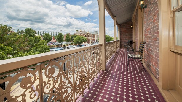 A balcony with patterned flooring, a decorative railing, and a view of a building and trees. - Cobb & Co Court Boutique Hotel (Mudgee)