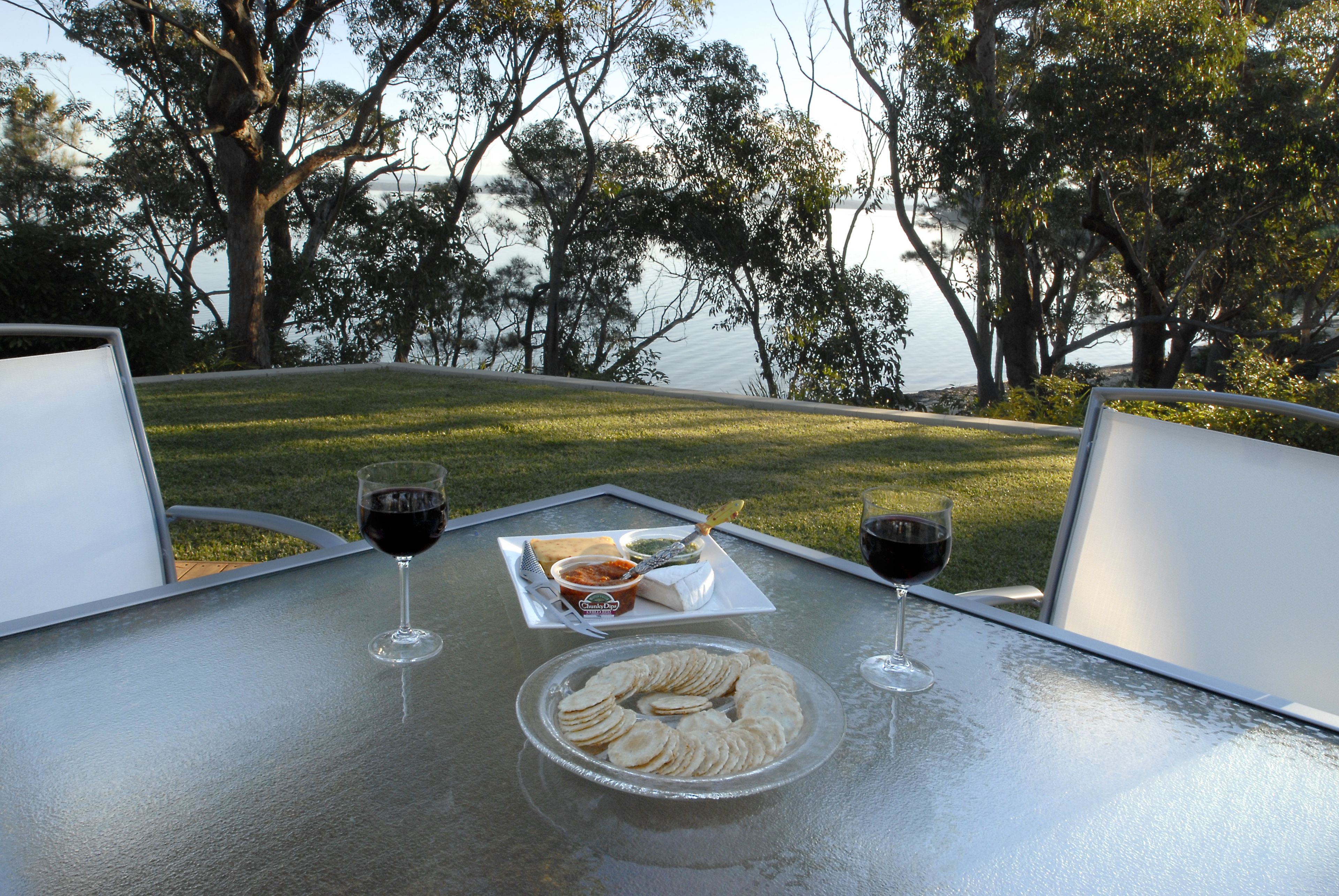 Une table avec un verre de vin rouge, une assiette de crackers et un petit bol de trempette, le tout placé devant un paysage verdoyant composé d’herbe et d’arbres.