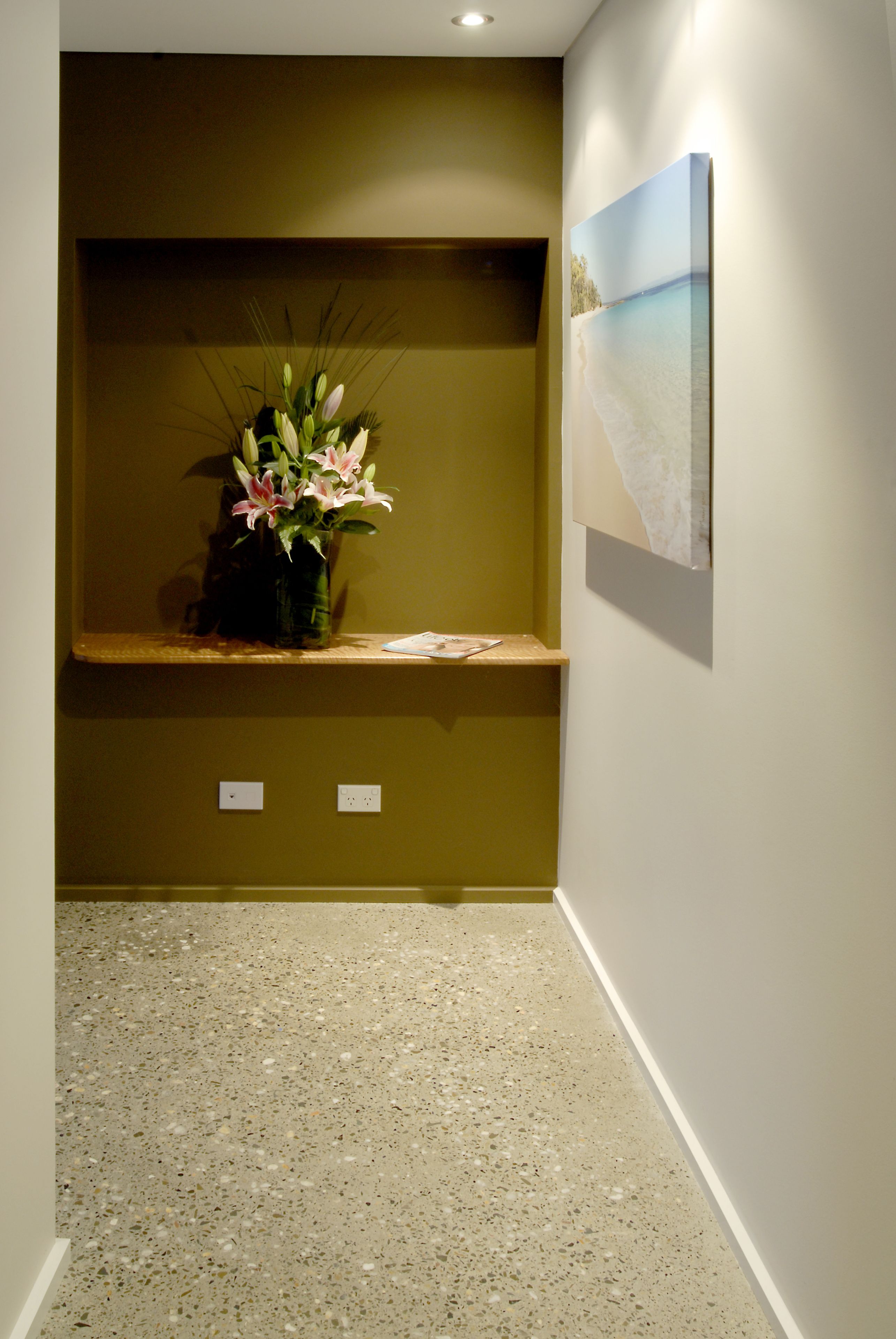 A hallway with a concrete floor, a shelf with a vase of flowers, and a painting on the wall.