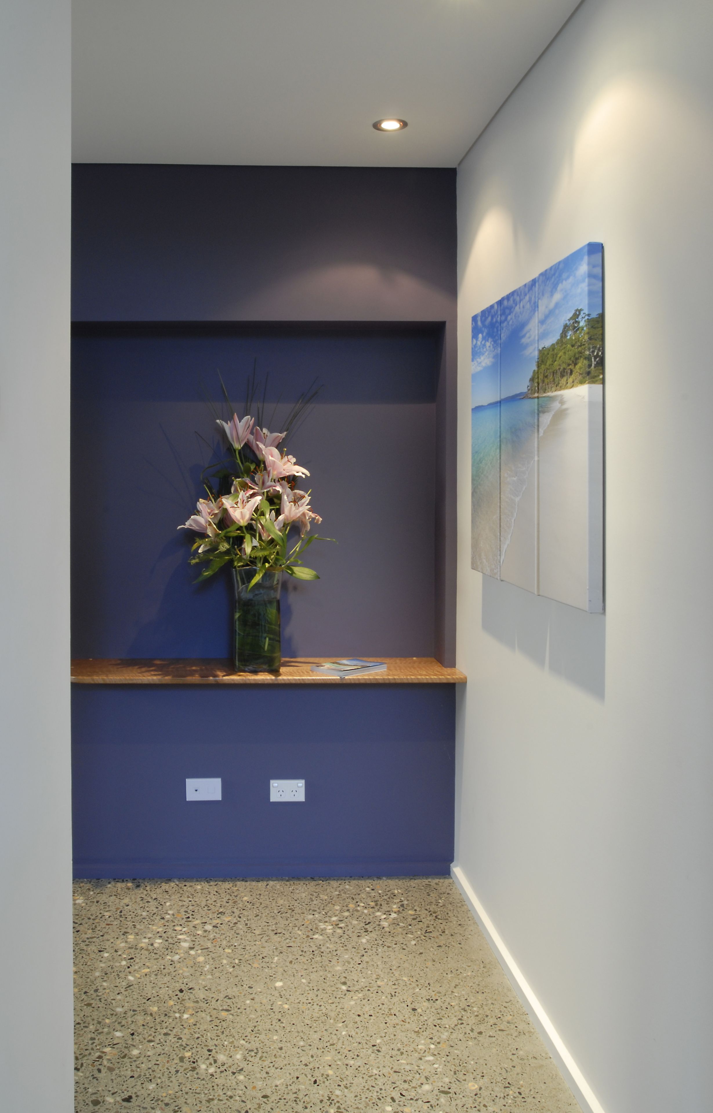 Ein Flur mit blauer Wand, einem Holzregal, einer Vase mit Blumen und einem Bilderrahmen mit Strandmotiv.