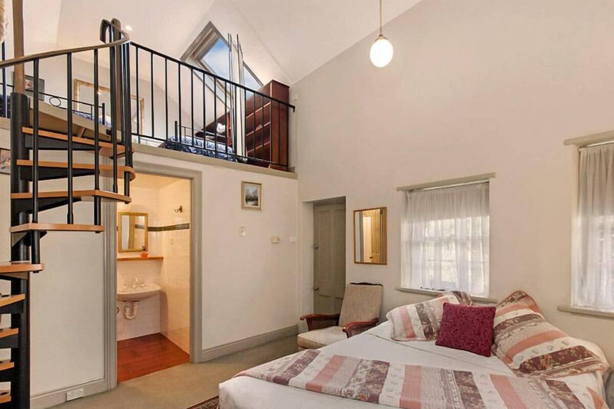 A bedroom with a large bed, a wooden staircase, a small bathroom, and a window with curtains.