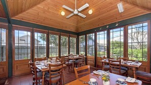 A spacious dining area with wooden tables and chairs, large windows, and a ceiling fan. - Victoria House (Portland)
