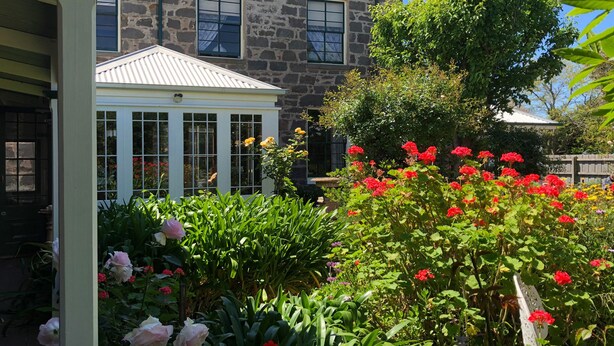 A stone house with a conservatory, surrounded by a garden with various flowers and a wooden fence. - Victoria House (Portland)