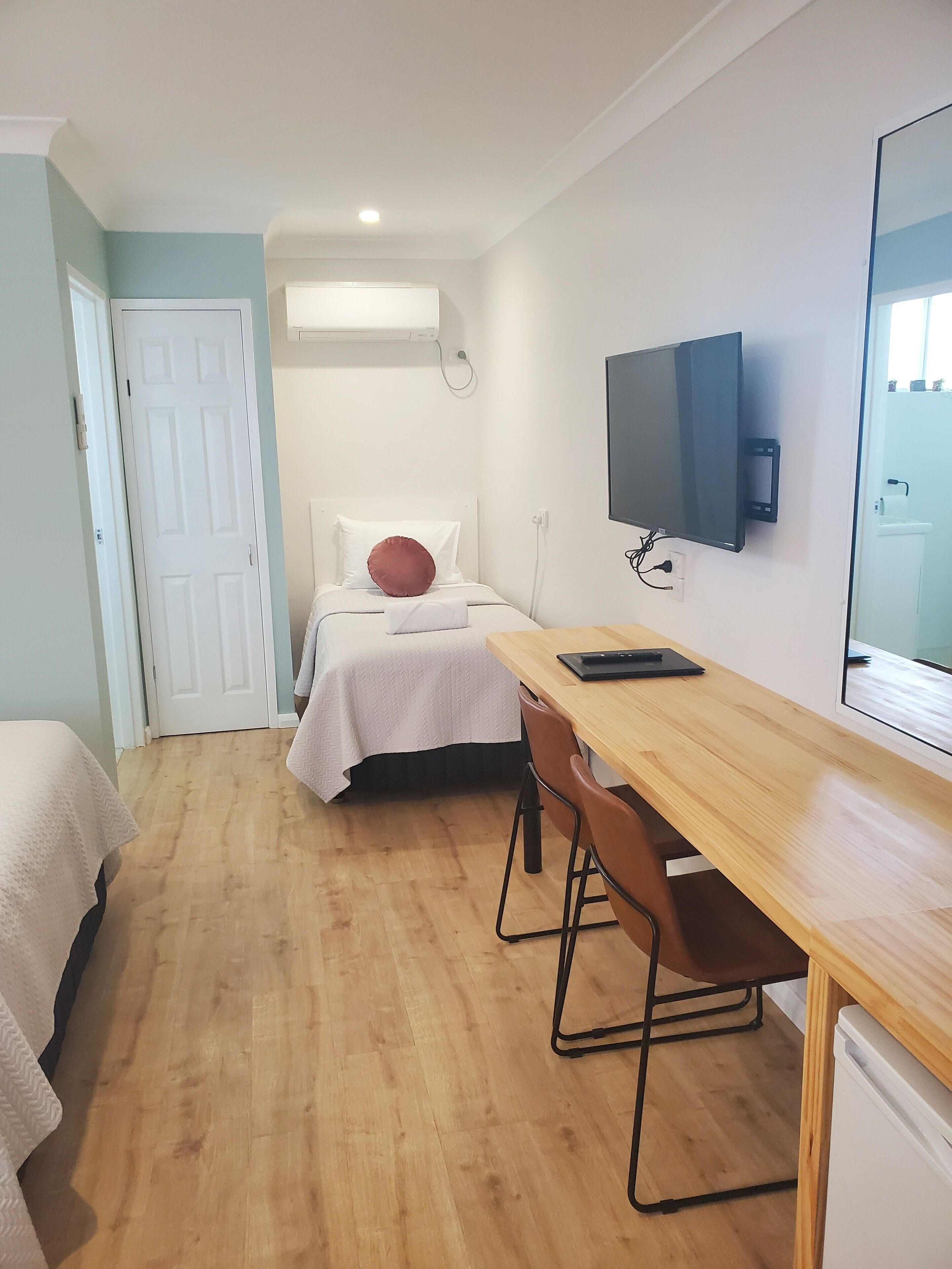 Een kleine, goed verlichte kamer met een bed, een bureau en een aan de muur bevestigde televisie.